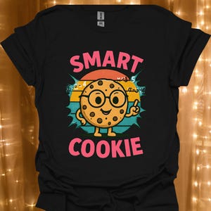 Può includere: T-shirt nero con una grafica colorata di un biscotto sorridente con gli occhiali e il pollice in su. Il testo "SMART COOKIE" è sopra e sotto l'illustrazione del biscotto, con equazioni matematiche sullo sfondo.