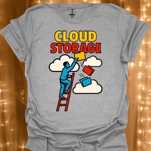 Può includere: Una t-shirt grigio melange con una grafica che mostra una persona che sale su una scala rossa verso nuvole bianche. Il testo CLOUD STORAGE è stampato in giallo e rosso. La persona tiene in mano una cartella gialla, mentre cartelle rosse e blu fluttuano.