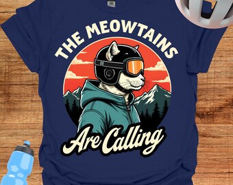 May include: Navy blue t-shirt featuring a cat wearing a ski helmet and goggles, with the text "The Meowtains Are Calling." The design includes a mountain scene and a sunset background. A blue water bottle and a white helmet are visible.