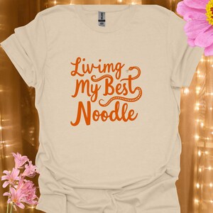 Op de afbeelding: Een beige t-shirt met korte mouwen en de oranje tekst "Living my Best Noodle" op de voorzijde gedrukt. Het ontwerp bevat een gestileerde oranje slang die in de letters is verwerkt, met een ronde hals en een standaard pasvorm.