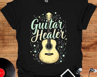 May include: Black t-shirt featuring a cream-colored acoustic guitar illustration with the words "Guitar Healer" in a whimsical font. The design includes musical notes and stars, creating a musical theme.