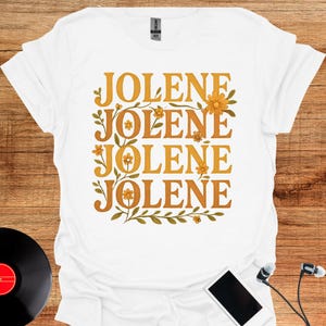 May include: A white short-sleeved t-shirt featuring the text JOLENE repeated four times in a stacked, golden-yellow serif font. The text is surrounded by illustrated green vines and small orange flowers, creating a decorative rectangular frame.