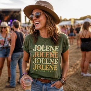 May include: An olive green short-sleeve t-shirt featuring the word JOLENE printed four times in a stacked, retro-style font with small floral illustrations integrated into the lettering, worn with a brown leather belt and layered bracelets.