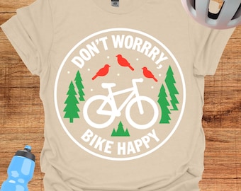Könnte beinhalten: Beiges T-Shirt mit einem kreisförmigen Aufdruck mit der Aufschrift "DON'T WORRRRY, BIKE HAPPY". Die Grafik zeigt ein weißes Fahrrad, rote Vögel und grüne Bäume. Eine blaue Wasserflasche und ein weißer Fahrradhelm sind ebenfalls zu sehen.