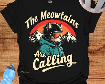 May include: Black t-shirt with a graphic of a cat wearing a ski helmet and goggles, with the text "The Meowtains Are Calling." The design includes a mountain range and a sunset.