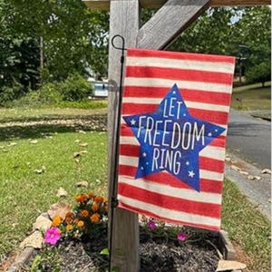 May include: A red and white striped garden flag with a blue star in the center. The star has the words "Let Freedom Ring" in white. The flag is hanging on a wooden post in a garden.