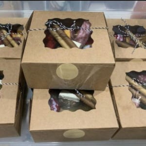 May include: Several small, brown cardboard boxes with a clear window revealing a variety of snacks. Each box is tied with twine and has a gold sticker. The boxes are stacked and arranged, suggesting a food gift.