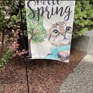 May include: A garden flag featuring a tabby cat with a blue bow, set against a spring scene with a flowering tree. The flag reads "Hello Spring" in elegant script, perfect for outdoor decor.