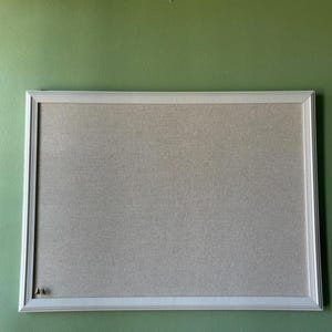 May include: A rectangular bulletin board with a white frame and a neutral-colored fabric surface. The board is mounted on a green wall. Two push pins are visible in the lower left corner. Ideal for displaying notes, photos, or reminders.