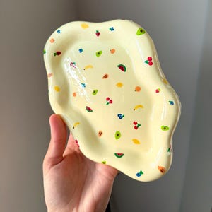 May include: A hand-held, irregular-shaped, yellow ceramic dish decorated with colorful fruit illustrations. The fruit designs include cherries, watermelon, bananas, and avocados. The dish has a glossy finish.