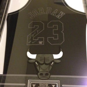 以下が含まれることがあります： 黒いフレームに入ったシカゴ・ブルズのジャージ。名前は「JORDAN」、背番号は「23」でグレー。サインとチームのブルロゴのグラフィックが含まれています。ジャージは黒いフレームに飾られています。