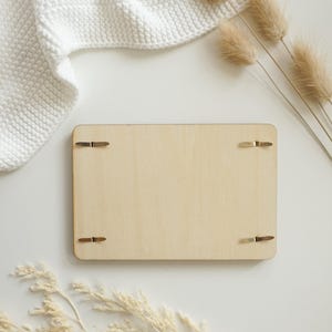 Puede incluir: Una tabla rectangular de madera de color claro con cuatro clips met&aacute;licos. La tabla est&aacute; rodeada por una manta blanca de punto, flores beige secas y una superficie blanca. La est&eacute;tica general es minimalista y natural.
