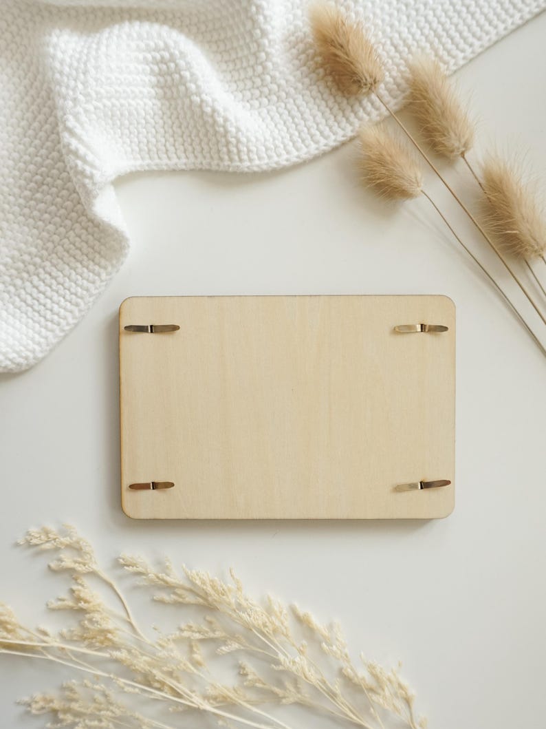 Puede incluir: Una tabla rectangular de madera de color claro con cuatro clips met&aacute;licos. La tabla est&aacute; rodeada por una manta blanca de punto, flores beige secas y una superficie blanca. La est&eacute;tica general es minimalista y natural.