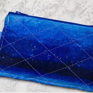 Pode incluir: Uma bolsa retangular acolchoada com um design gradiente azul, com costuras brancas em padrão de diamante. A bolsa tem um zíper azul e um pequeno laço azul com um anel de metal preso. O tema geral é um design celestial ou de céu noturno.