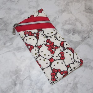 Könnte beinhalten: Ein rechteckiges, gestepptes Federmäppchen mit einem sich wiederholenden Muster von Hello Kitty-Gesichtern mit roten Schleifen auf weißem Grund. Der obere Teil ist rot mit einem silbernen Reißverschluss.