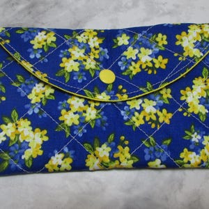 May include: A quilted clutch with a blue background and a floral pattern in yellow and white. The clutch has a curved flap with a yellow button closure and yellow trim. The stitching is white.