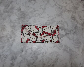 May include: A white wallet with a red trim and a repeating pattern of Hello Kitty faces with red bows. The wallet has a flap closure with a black button.