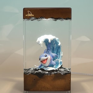 May include: A decorative lamp featuring a cartoon shark emerging from a blue wave. The lamp has a rectangular wooden base and a clear acrylic enclosure. The text "Joey's Adventures" is visible on the top.