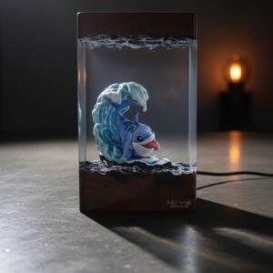 May include: A whimsical resin art piece featuring a cartoon shark emerging from a blue wave. The shark has a red tongue and is set within a clear rectangular block with a wooden base. The piece is lit, creating a playful and unique decorative item.