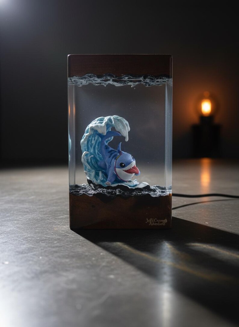 May include: A whimsical resin art piece featuring a cartoon shark emerging from a blue wave. The shark has a red tongue and is set within a clear rectangular block with a wooden base. The piece is lit, creating a playful and unique decorative item.