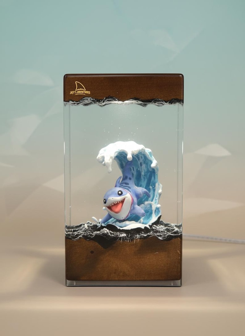 May include: A decorative lamp featuring a cartoon shark emerging from a blue wave. The lamp has a rectangular wooden base and a clear acrylic enclosure. The text "Joey's Adventures" is visible on the top.