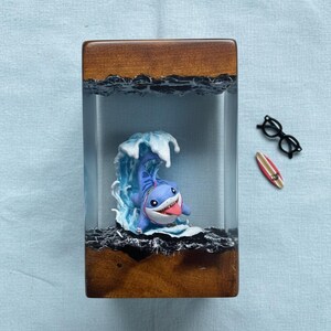 May include: A wooden and resin art piece featuring a cartoon shark surfing a wave. The shark is blue with a white belly and a red tongue. The wave is white and blue. A small surfboard and glasses are next to the art piece.