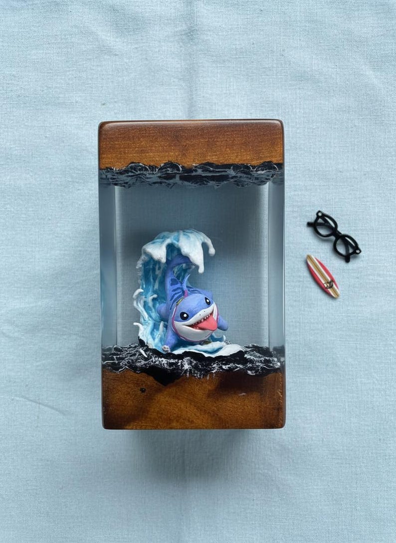 May include: A wooden and resin art piece featuring a cartoon shark surfing a wave. The shark is blue with a white belly and a red tongue. The wave is white and blue. A small surfboard and glasses are next to the art piece.