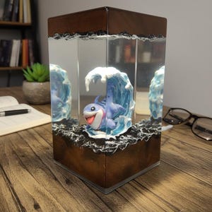 May include: A decorative lamp featuring a cartoon shark in a wave, encased in a clear rectangular block. The base and top are a dark brown wood. The shark is blue with a white belly and a red tongue. The lamp sits on a wooden surface.