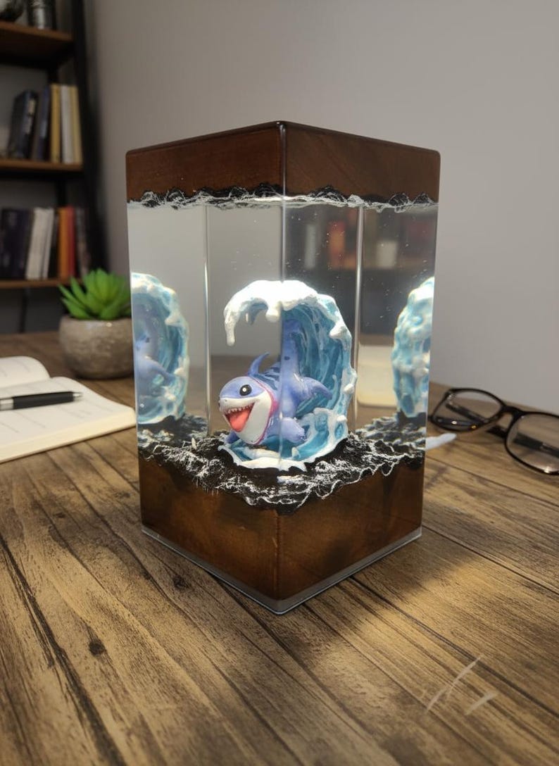 May include: A decorative lamp featuring a cartoon shark in a wave, encased in a clear rectangular block. The base and top are a dark brown wood. The shark is blue with a white belly and a red tongue. The lamp sits on a wooden surface.
