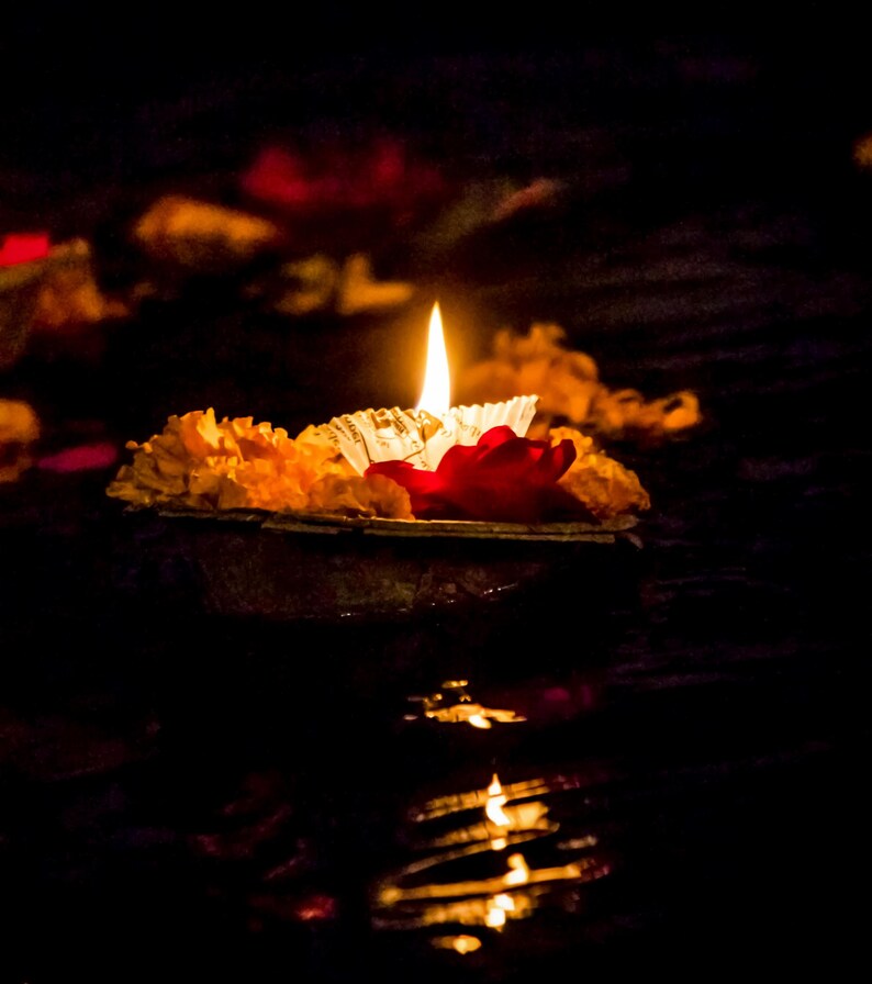 May include: A lit candle floats on water, surrounded by orange marigold flowers and a red petal. The flame casts a warm glow against the dark background, creating a serene and spiritual ambiance.