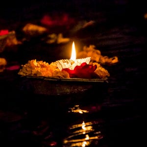 Pode incluir: Uma vela acesa flutua na &aacute;gua, rodeada por flores de cal&ecirc;ndula laranja e uma p&eacute;tala vermelha. O fundo escuro real&ccedil;a o brilho da chama, com reflexos na &aacute;gua.
