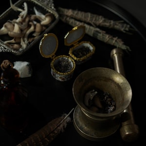 May include: A collection of items on a black surface, including small decorative boxes, a mortar and pestle, dried herbs, and a feather. The boxes are open, revealing their contents. The scene has a mystical or apothecary theme.