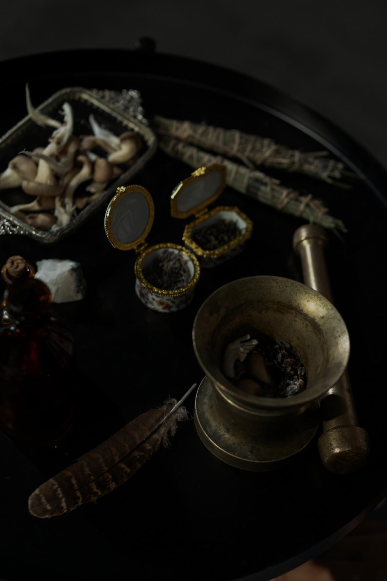 May include: A collection of items on a black surface, including small decorative boxes, a mortar and pestle, dried herbs, and a feather. The boxes are open, revealing their contents. The scene has a mystical or apothecary theme.