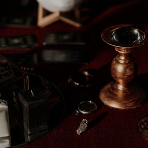 May include: A still-life image featuring a collection of mystical items. A gold-colored candle holder with a clear glass orb sits next to a purple candle. A small glass bottle with a cork stopper and a clear liquid is visible, along with a small crystal.