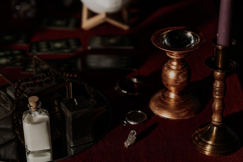 May include: A still-life image featuring a collection of mystical items. A gold-colored candle holder with a clear glass orb sits next to a purple candle. A small glass bottle with a cork stopper and a clear liquid is visible, along with a small crystal.