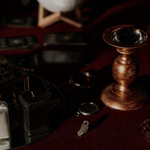 Pode incluir: Uma natureza morta com est&eacute;tica vintage. A cena inclui um casti&ccedil;al dourado, uma pequena garrafa de vidro com rolha e uma vela roxa. Outros itens incluem um cristal e uma bandeja espelhada.