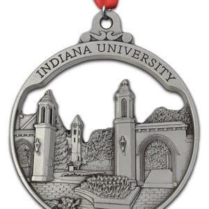 May include: A silver-colored Indiana University ornament with a red ribbon. The ornament features a detailed depiction of a university building and the words "INDIANA UNIVERSITY" in a circular design.