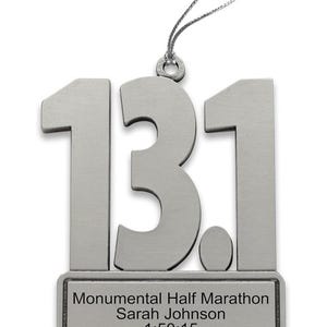 May include: Silver-colored metal ornament in the shape of "13.1" with a small rectangular plaque below. The plaque reads "Monumental Half Marathon Sarah Johnson 1:50:15". A silver string is attached for hanging.