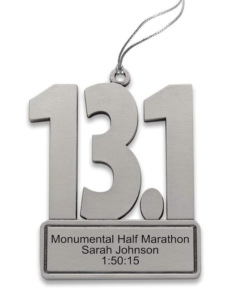 May include: Silver-colored metal ornament in the shape of "13.1" with a small rectangular plaque below. The plaque reads "Monumental Half Marathon Sarah Johnson 1:50:15". A silver string is attached for hanging.