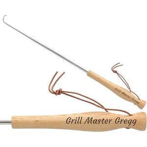 May include: A stainless steel grill hook with a wooden handle. The handle is engraved with "Grill Master Gregg" and has a leather loop for hanging. The hook is designed for grilling and barbecue use.