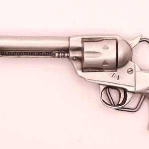 May include: A silver-toned metal belt buckle shaped like a classic revolver. The design features a long barrel, a rotating cylinder, a trigger guard, and a textured handle grip. The metallic finish has a smooth sheen with visible screw details.