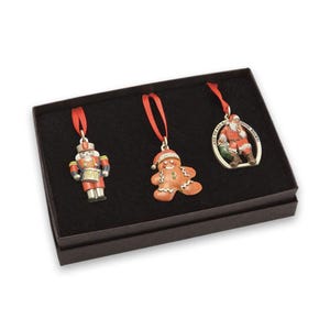 May include: A set of three Christmas ornaments in a black box, featuring a nutcracker in a red and blue uniform, a brown gingerbread person, and a circular metal frame with a Santa Claus figure and the text AND TO ALL A GOOD NIGHT.