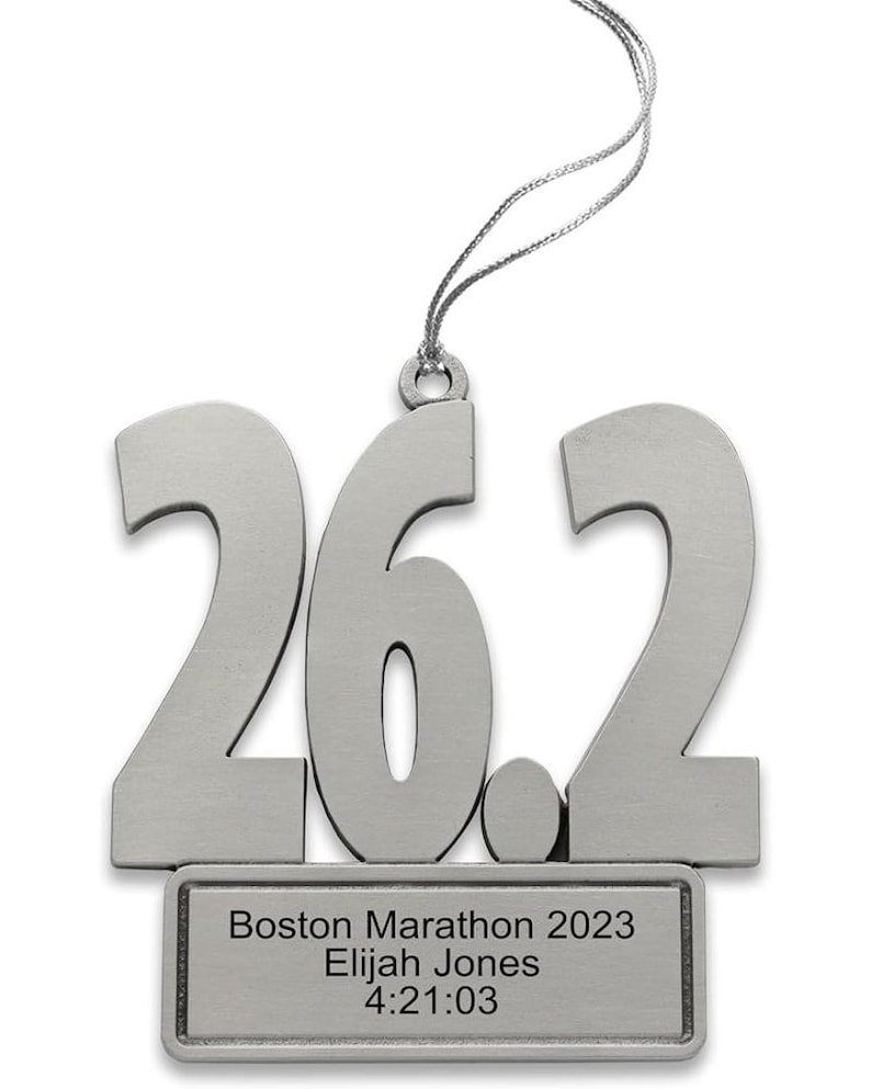 May include: A silver-colored ornament in the shape of the numbers "26.2", representing the marathon distance. Below the numbers is a rectangular plaque with the text "Boston Marathon 2023 Elijah Jones 4:21:03". A silver string is attached for hanging.