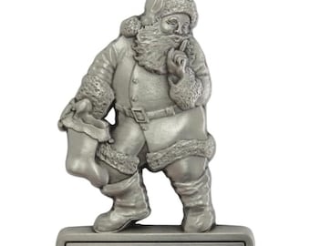 May include: A silver-colored Christmas ornament featuring Santa Claus with a finger to his lips, holding a stocking. The ornament has the text "The Davidson Family Merry Christmas 2023" on a rectangular base. The ornament is suspended by a silver cord.