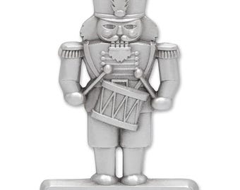 May include: Silver-colored Christmas ornament shaped like a toy soldier, holding drumsticks and a drum. The ornament has a rectangular base with the text "The Williams Family Merry Christmas 2023".
