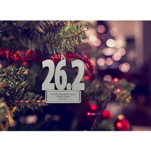 May include: A silver Christmas tree ornament with the number "26.2" cut out. Below the numbers is a rectangular plaque with the text "Boston Marathon 2023, Elijah Jones, 4:21:03". The ornament hangs on a Christmas tree.