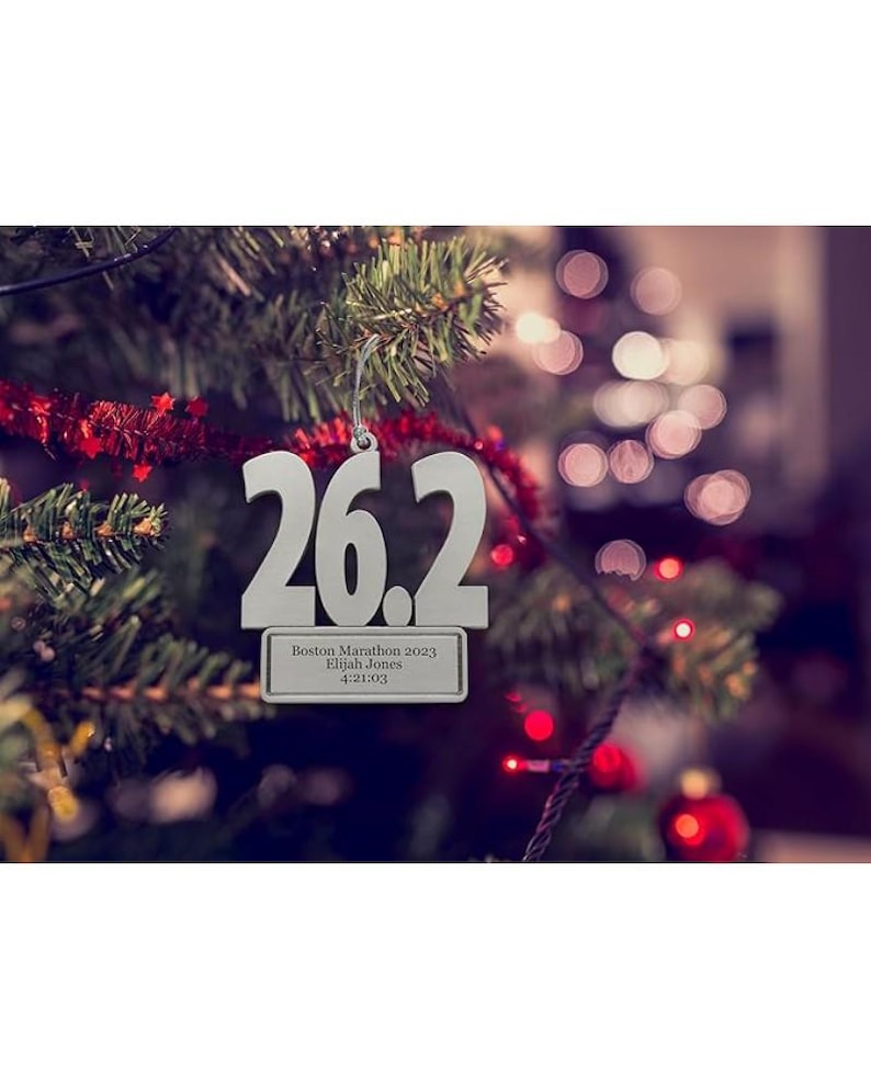 May include: A silver Christmas tree ornament with the number "26.2" cut out. Below the numbers is a rectangular plaque with the text "Boston Marathon 2023, Elijah Jones, 4:21:03". The ornament hangs on a Christmas tree.