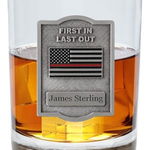May include: A clear rocks glass filled with amber liquid and ice cubes. A silver plaque with the text "FIRST IN LAST OUT" and a US flag with a red stripe is attached to the glass. The name "James Sterling" is below the flag.