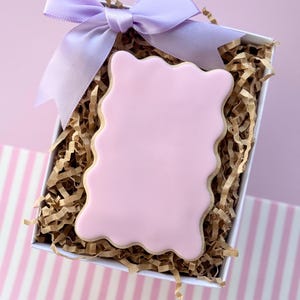Könnte beinhalten: Ein mit Rosa und Gold verzierter Keks in einer weißen Geschenkbox mit braunem zerkleinertem Papier und einer lila Schleife.