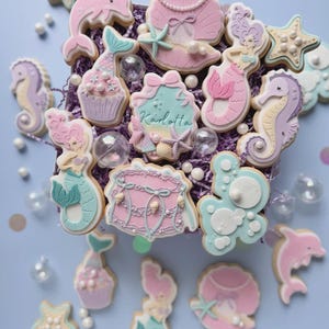 Könnte beinhalten: Eine Auswahl an Keksen zum Thema Meerjungfrau in Pastellfarben. Die Kekse zeigen Formen von Meerjungfrauen, Muscheln, Delfinen, Seepferdchen und Cupcakes. Einige Kekse tragen den Namen "Karlotta". Die Kekse sind aufwendig verziert und von dekorativen Elementen umgeben.